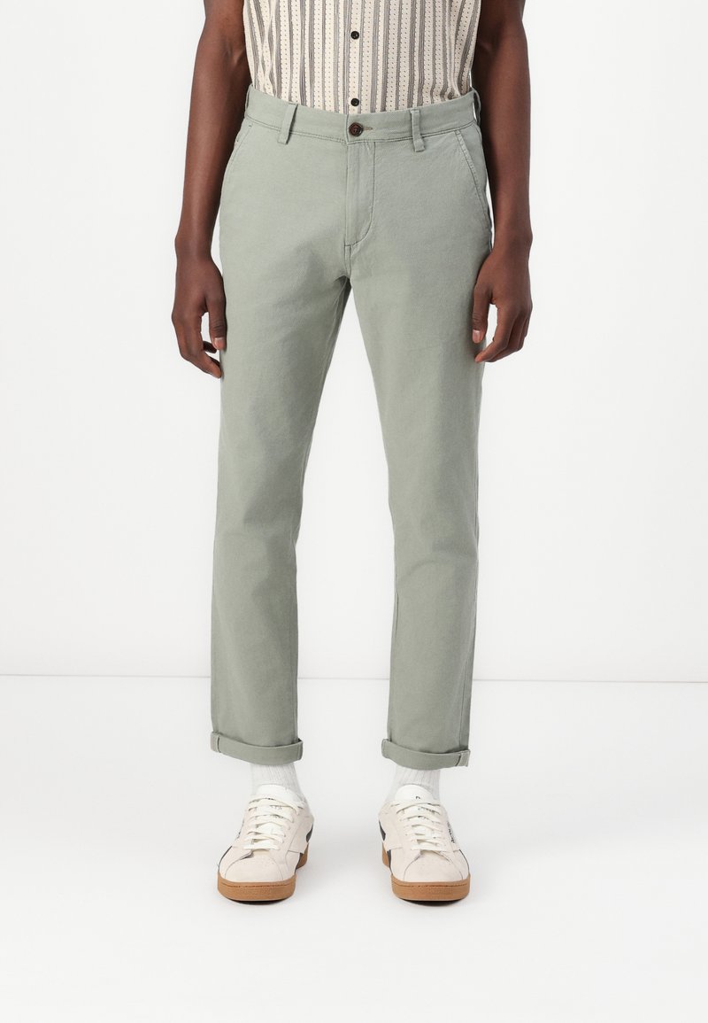 Pantalon en coton vert clair avec une coupe droite, présentant une fermeture à bouton, des poches avant et des ourlets roulés. Associé à des baskets blanches.