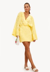 Vestido mini amarillo con patrón floral texturizado, escote en V profundo y mangas acampanadas. Combinado con zapatos beige de punta abierta y un pequeño bolso blanco.