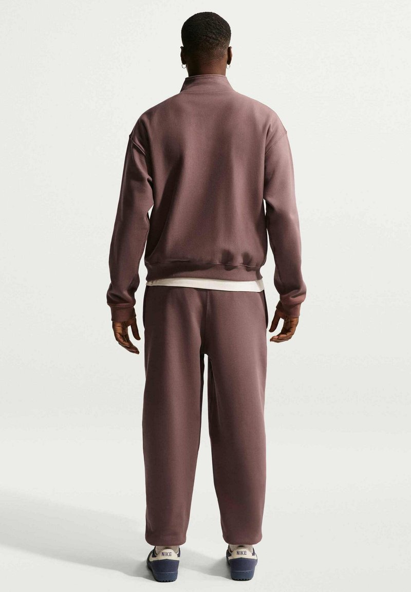 Man staat met zijn rug naar de camera, draagt een mauvekleurig trainingspak en witte Nike-sneakers tegen een effen witte achtergrond.