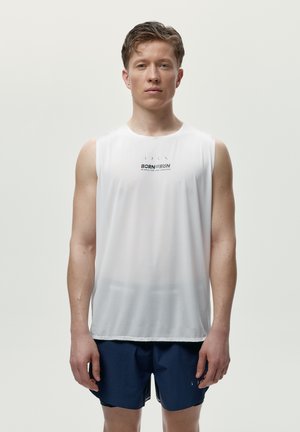 Junger Mann mit kurzen Haaren, der ein weißes ärmelloses Sporthemd und dunkelblaue Laufshorts trägt, steht vor einem schlichten Hintergrund.
