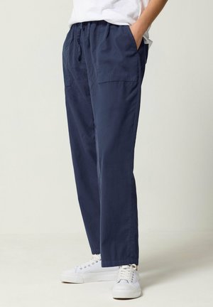 Persoon draagt marineblauwe casual broek met elastische taille en witte sneakers, staat met handen in de zakken tegen een effen achtergrond.