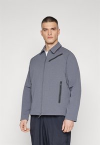 Gray zip-up jacket with a textured fabric, featuring a stand collar and side pockets, paired with a dark bottom garment.