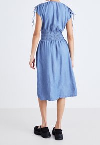 Nainen, jolla on päällään vaaleansininen midi-pitkä farkkuasu, jossa on kerätty vyötärö ja lyhyet solmittavat hihat, seisoo selkä kameraan päin mustissa lenkkareissa.