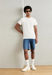T-shirt en coton blanc, shorts en denim bleu, baskets blanches avec des accents beiges, et lunettes de soleil vertes. Modèle posé contre un mur neutre.