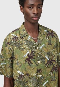 Κοντομάνικο πουκάμισο από πράσινο ύφασμα με τροπικό σχέδιο φύλλων που περιλαμβάνει παλμιές και κείμενα. Λευκά κουμπιά και σχεδίαση με άνετη εφαρμογή.