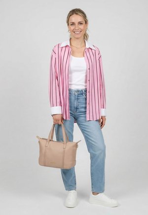 Vrouw glimlachend, gekleed in een roze gestreept shirt, wit topje, blauwe spijkerbroek, witte sneakers, met een beige handtas, tegen een effen achtergrond.