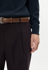 Tengerészkék pulóver, barna bőöv ezüst csattal, és sötét nadrág, amelyen gyűrődés és oldalsó zseb található. Közeli felvétel a derékról és a kézről.