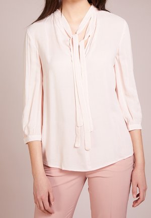 Nainen pukeutuneena vaaleanpunaiseen puseroon, jossa on solmittava kaulanympärystys ja kolme neljäsosahihasta, yhdistettynä vastaaviin vaaleanpunaisiin housuihin.