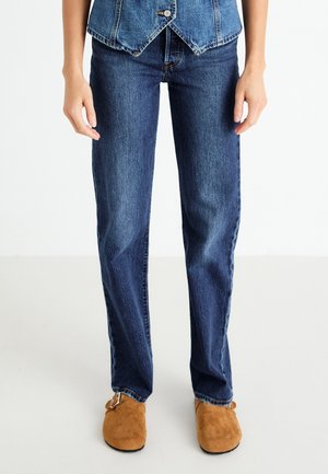 Persoon die recht model blauwe spijkerbroek draagt, een blauw denim vest en bruine suède instapschoenen met gespen.