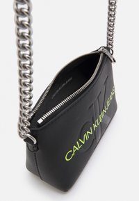 Borsa a mano in pelle nera con tracolla a catena d'argento, logo in rilievo audace e marchio verde neon sul davanti. Apertura con cerniera visibile.