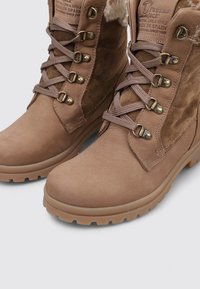 Botas de tobillo de cuero tostado con un acabado texturizado, que presentan herrajes metálicos, cordones reforzados y una suela de goma gruesa. Detalle de forro de pelaje sintético.