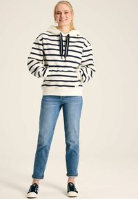 Pruhovaná mikina v krémovej a námorníckej modrej farbe, s kapucňou na šnúrku, klokánkovými vreckami a rebrovanými manžetami, kombinovaná so svetlými džínsami a tmavými topánkami.