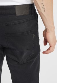 Crne traperice imaju stražnji džep s pravokutnom zakrpom. Tekstura je glatka, a kroj izgleda usko i prilagođeno.