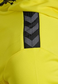 Gul atletisk stof med en tekstureret overflade, der har et sort sidelæns panel med et chevron-mønster og reflekterende detaljer. Justerbar snor.