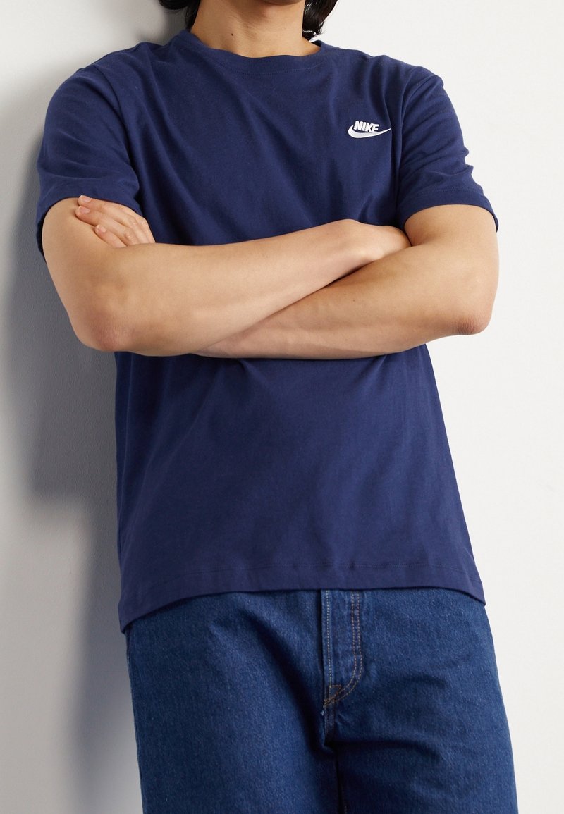 Mörkblå bomullströja med korta ärmar, med en vit Nike-logotyp på vänster bröstområde. Matchas med blå jeans.