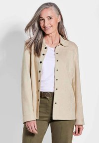 Beige suède jack met zwarte drukknoopjes, een kraag en lange mouwen, gedragen over een witte top en olijfgroene broek.