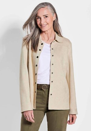 Veste en daim beige avec des boutons-pression noirs, un col, et des manches longues, portée par-dessus un haut blanc et un pantalon vert olive.