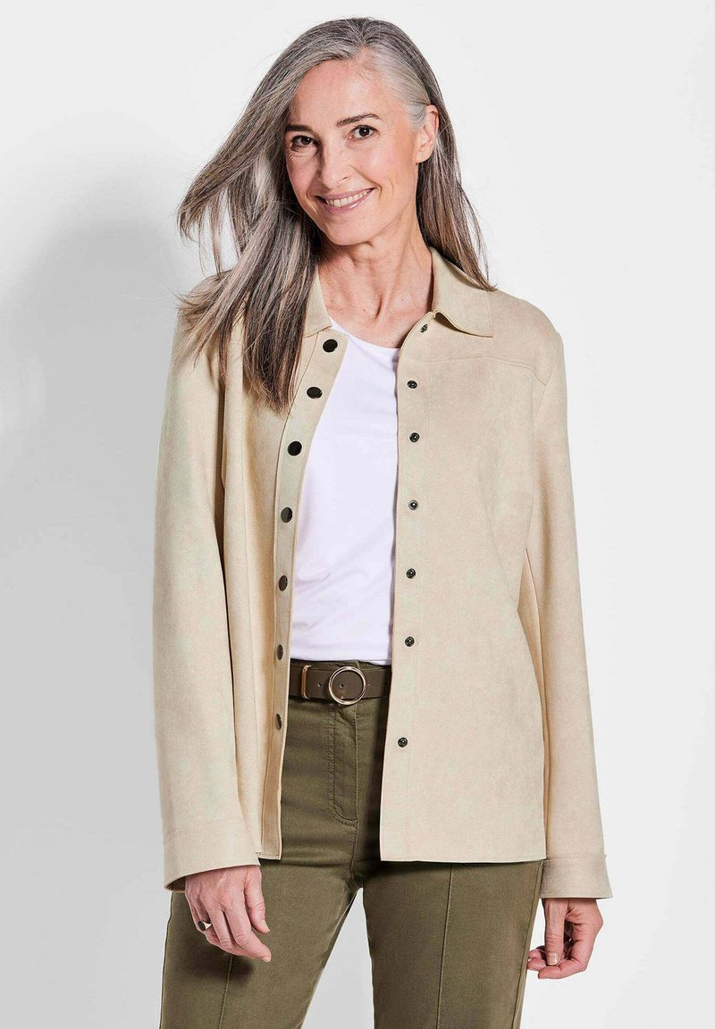 Beige suède jack met zwarte drukknoopjes, een kraag en lange mouwen, gedragen over een witte top en olijfgroene broek.