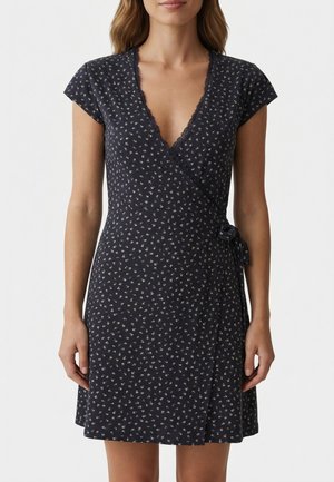 Mujer vistiendo un vestido cruzado negro con flores, de mangas cortas y detalles de encaje, ceñido en la cintura, sobre un fondo liso.