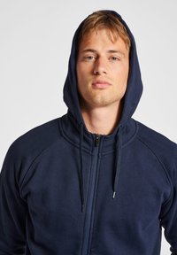 Felpa con zip di colore blu navy, dotata di un ampio tascone frontale, cappuccio con cordino regolabile e una texture liscia e morbida; presenta maniche raglan e un design aderente.