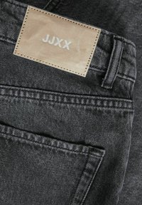Jeans de mezclilla negra con una etiqueta de cuero visible etiquetada como "JJXX", con detalles de costura y un bolsillo trasero. El material parece duradero y texturizado.