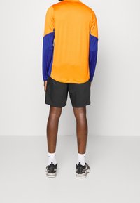 Kortærmet sportstrøje med orange overdel og blå ærmer, parret med sorte shorts. Modellen har grå sneakers og hvide sokker på.