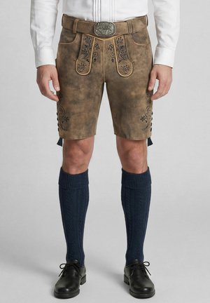 Mann trägt traditionelle braune Lederhosen mit Stickerei, marineblaue gestrickte knielange Socken, schwarze Schuhe und ein weißes Hemd.
