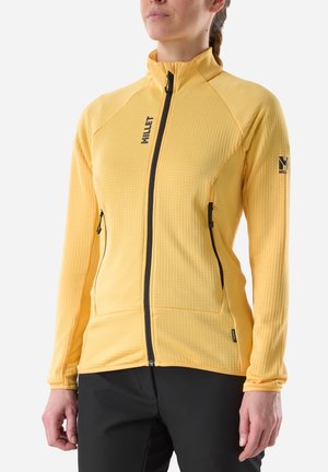 Woman wearing a yellow zip-up jacket with black zippers, two front pockets, and Millet logos on chest and left sleeve.
