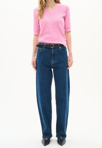 Kvinna i en rosa kortärmad V-ringad topp, högmidjade blå jeans med ett svart bälte, och svarta spetsiga skor som står mot en vit bakgrund.