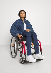 Denim jakna s napihnjenimi rokavi, bela majica in modri kavbojke. Bele teniske. Sedeči v rdečem in srebrnem invalidskem vozičku na nevtralni podlagi.