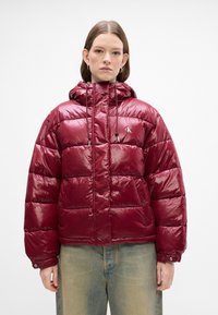 Kvinne iført en skinnende kastanjefarget quiltet hettejakke med frontlommer og snøring, kombinert med lyseblå jeans, står vendt mot kamera.