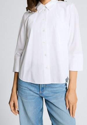 Femme portant une chemise blanche boutonnée avec des manches trois-quarts et un jean en denim bleu clair sur un fond uni.