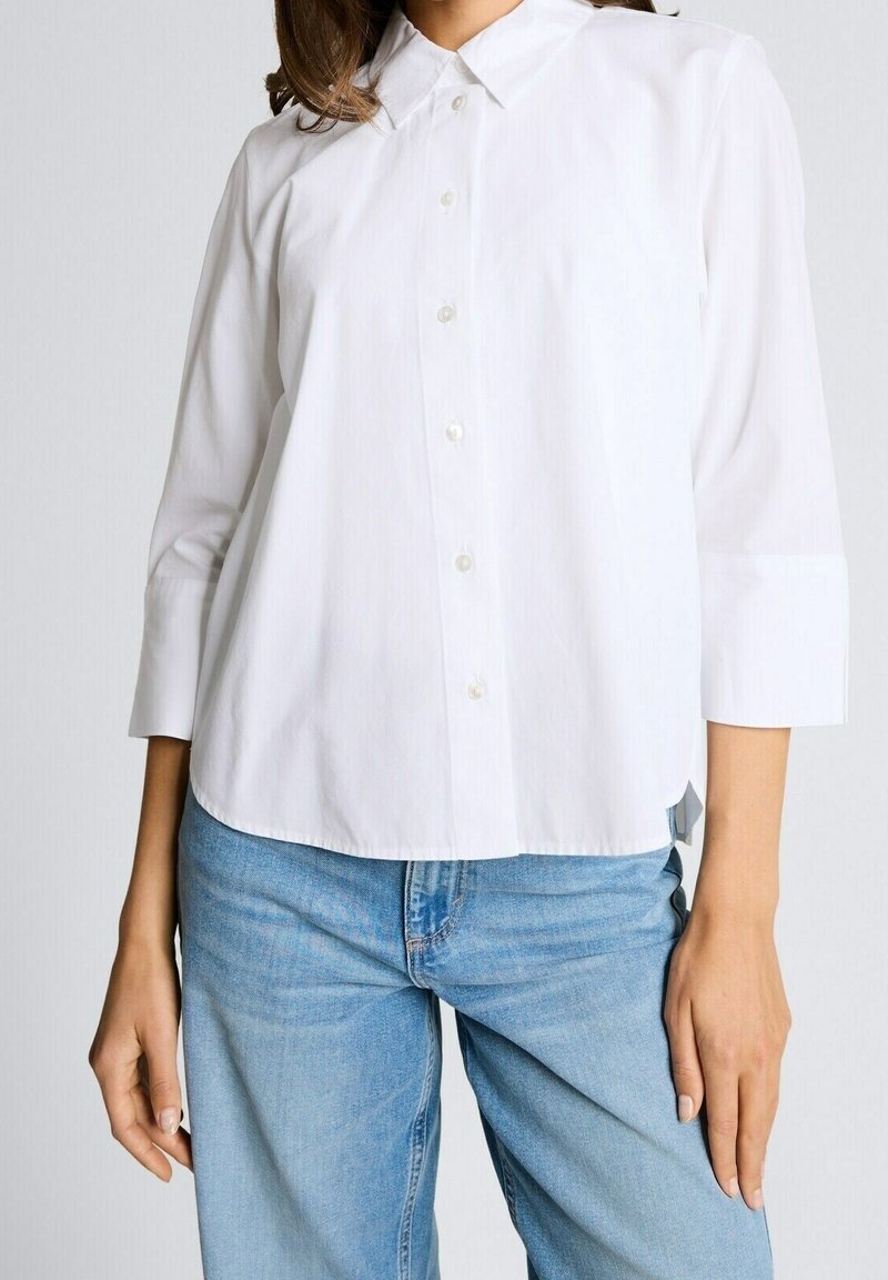 Femme portant une chemise blanche boutonnée avec des manches trois-quarts et un jean en denim bleu clair sur un fond uni.