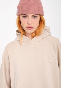 Beige hooded sweatshirt with a relaxed fit, featuring a small white logo patch and soft texture. Pink knitted beanie completes the look.