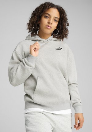 Ung kvinde med krøllet hår iført en lys grå Puma-hoodie og matchende bukser, som holder fast i hoodie-snorene foran en ensfarvet grå baggrund.