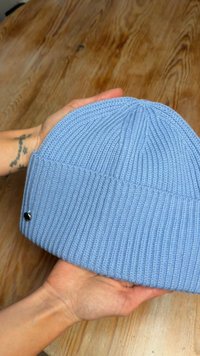 Világoskék kötött sapka bordázott textúrával és hajtott szegéllyel. Az oldalon egy kis fekete díszítő elem található.