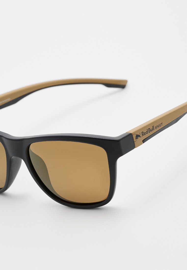 Czarno-brązowe okulary przeciwsłoneczne z kanciastymi oprawkami i brązowymi soczewkami. Posiadają logo Red Bull na zauszniku. Gładki materiał plastikowy.