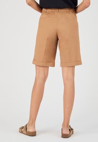 Pantalones cortos marrones, ajustados, con dobladillo enrollado, cinturilla elástica y dos bolsillos traseros. Llevados con sandalias marrones de tipo slide, sobre un fondo blanco.