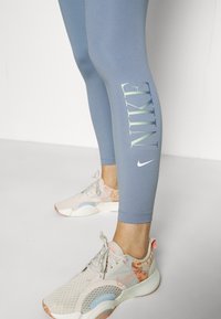 Leggings azzurri con la scritta "Nike" stampata verticalmente. Abbinati a sneakers beige con accenti rosa e blu, progettate per l'uso atletico.