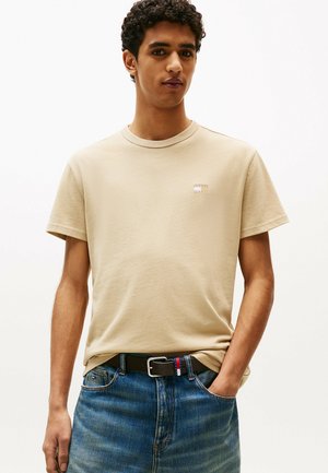 T-shirt in cotone beige con scollatura rotonda, maniche corte e un piccolo logo quadrato sul petto. Indossato con jeans in denim blu.
