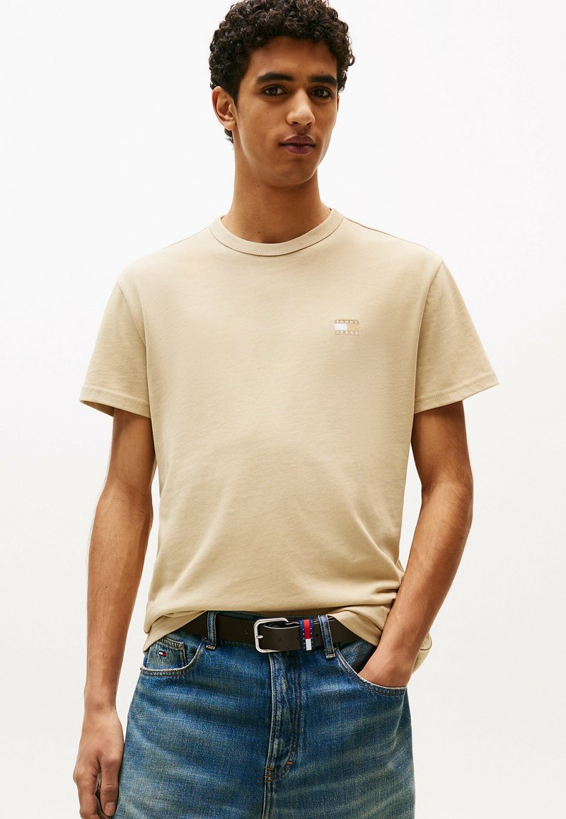 T-shirt in cotone beige con scollatura rotonda, maniche corte e un piccolo logo quadrato sul petto. Indossato con jeans in denim blu.