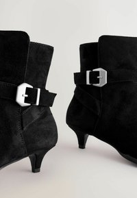 Bottines en suede noir avec un bout pointu, un petit talon et un accent en boucle en ton argent sur le côté, présentant des détails de couture contrastés.