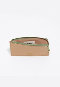 Cartera de cuero beige con un zipper verde, herrajes dorados y una etiqueta interior. Diseño compacto, forma simple, texturas sutiles.