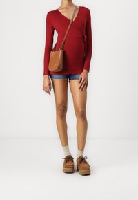 Rote langärmlige Wickeltop mit Seitenschnürung, kombiniert mit Denim-Shorts. Braune Umhängetasche, hellbraune Plateauschuhe und beige Socken.