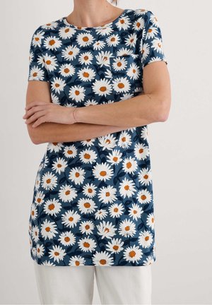 Kvinde iført en mørkeblå kortærmet skjorte med hvidt tusindfryd blomsterprint og hvide bukser, arme krydset over brystet.