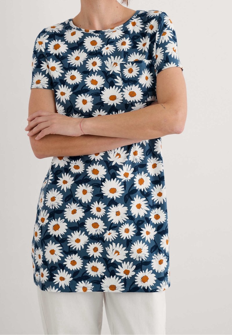 Femme portant une chemise à manches courtes bleu marine avec un motif floral de marguerites blanches et un pantalon blanc, les bras croisés sur la poitrine.
