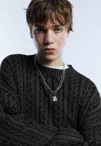 Čierny pletený sveter s vystúpenými vzormi v podobe vrkočov, šedý kovový reťazový náhrdelník s detailným príveskom a vrstvená biela košeľa pod ním.