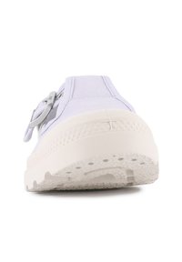 Zapatilla de lona blanca con suela de goma texturizada gruesa, hebilla metálica redonda en el costado, vista de frente sobre un fondo blanco.