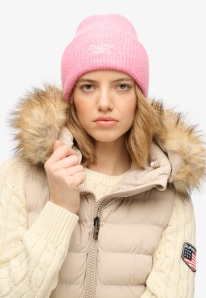 Joven con un gorro de punto rosa, suéter crema de punto trenzado y chaleco acolchado beige con capucha forrada de piel, sujetando el cuello cerca.