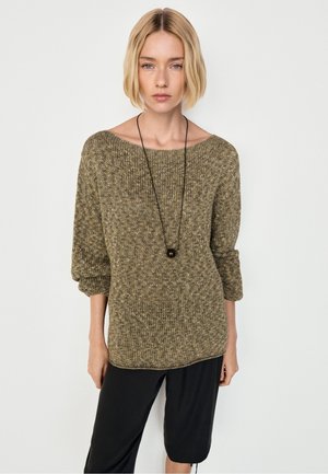 Blond meisje draagt een olijfgroene gebreide trui met opgerolde mouwen en een zwarte hangerketting, gecombineerd met een zwarte broek.